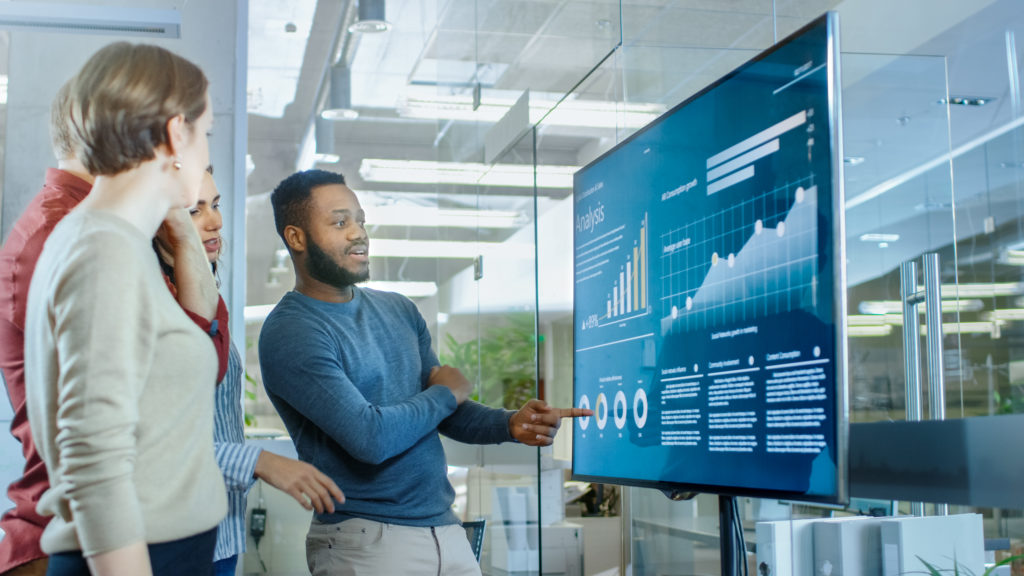 Diverse Team Of Young Professionals In Conference Room Have Discussion About Statistics And Graphs Shown On A Presentation Tv.